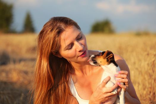 Young brunette woman with long hair, holding Jack Russell terrier puppy that is trying to lick her on face. Wheat field lit by afternoon sun in background.