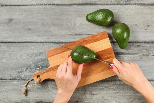 Tabletop view, woman hands holding chef knife, cutting avocado on chopping board, two whole avocados near on gray wood desk.