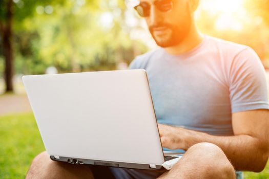 Young man using and typing laptop computer in summer grass. Freelancer working in outdoor park. Sun flare