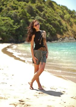 Young slim woman in black t shirt and shorts, wearing sunglasses, wind in her dark hair, standing on the beach with azure ocean in background. Koh Lanta, Thailand.