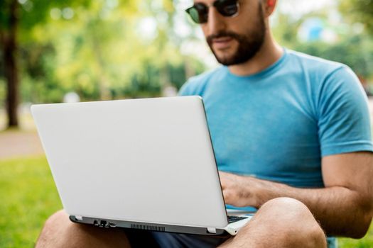 Young man using and typing laptop computer in summer grass. Freelancer working in outdoor park