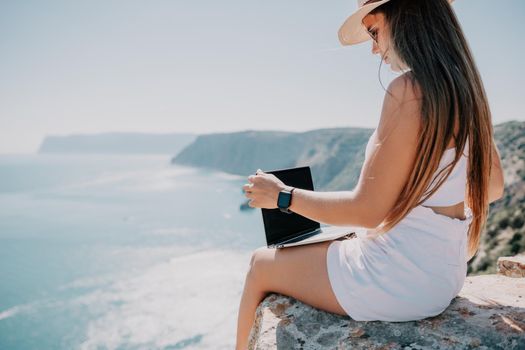 Successful business woman in yellow hat working on laptop by the sea. Pretty lady typing on computer at summer day outdoors. Freelance, travel and holidays concept.