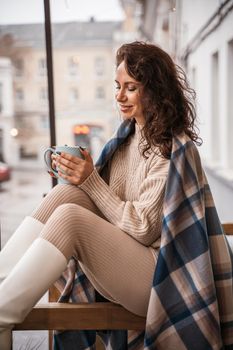 A middle-aged woman in a beige sweater with a blue mug in her hands is in a street cafe on the veranda.