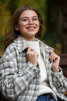 European elegant young woman in a stylish trench coat in a white sweater and blue jeans resting on the bench. warm autumn day. brown wooden bench