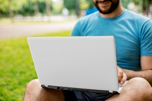 Young man using and typing laptop computer in summer grass. Freelancer working in outdoor park