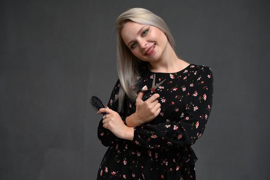 Young blondy woman over isolated gray background with hairdresser scissor and comb. Looking happy and smiling. Ready for haircut.