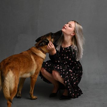 Adult girl carries dog which embraces her around neck walk together isolated over gray background. Female dog lover poses with pet looks away likes animals. Care and love