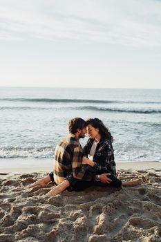 Young woman and man hugging while sitting together on the seashore with sea waves on background at sunrise