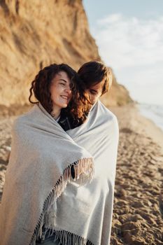 Young cheerful woman and man standing on the seashore wrapped in plaid at sunrise