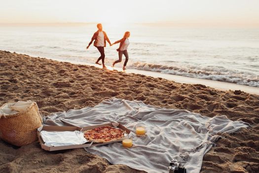 Focus on picnic set with pizza and juice, happy couple, woman and man holding by hands and running along the coastline together on background