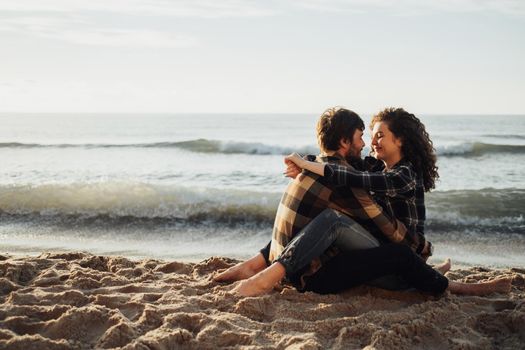 Young couple, curly haired woman with brunette man fall in love sitting and hugging on seashore at sunrise