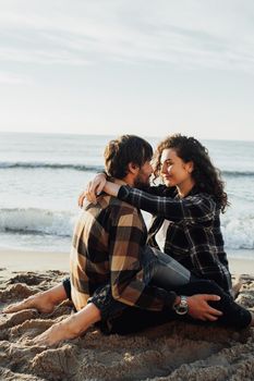 Young woman and man hugging while sitting together on the seashore with sea waves on background at sunrise