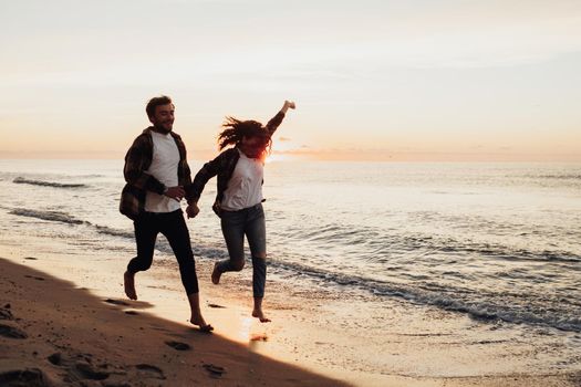 Happy young couple holding by hands and running along the coastline, woman and man meeting sunrise on sea together