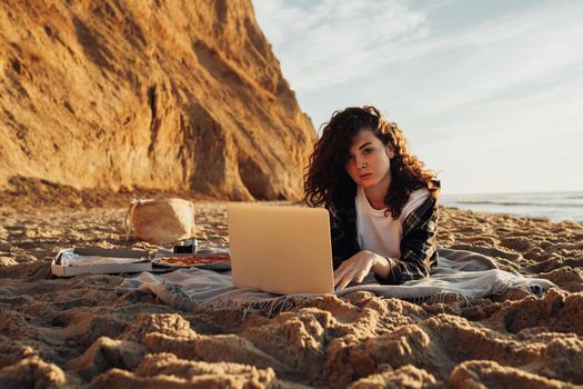 Young curly haired woman working on laptop while laying on seashore at dawn