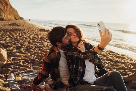 Happy young adult couple falling in love, curly haired woman making selfie together with boyfriend while sitting on the seashore at dawn