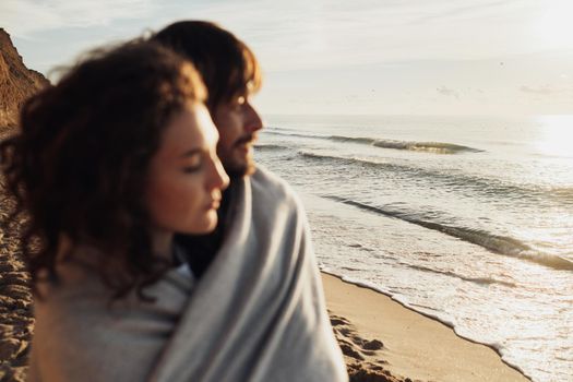 Blurred young adult couple wrapped in plaid and standing on the seashore, focus on sea waves