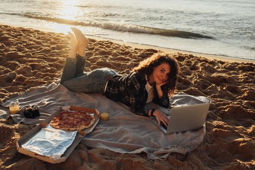 Happy curly haired woman eating pizza and working on laptop while laying on seashore at dawn