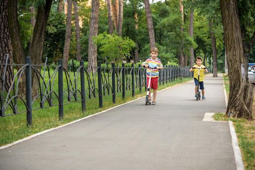 Two attractive European boys brothers, wearing red and white checkered shirts, standing on scooters in the park. They laughing, smiling, hugging and having fun. Active leisure time with kids.