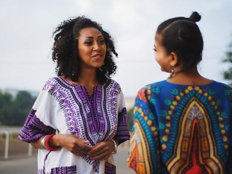 Two happy beautiful afro-american women friends walking outdoor. Young mixed race girls wearing colorful clothing communicates, enjoys the meeting and laughing