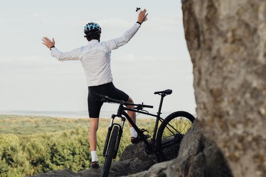 Equipped Professional Male Cyclist Sitting with Mountain Bike on Rock, Adult Man Raising Hands and Enjoying His Extreme Hobby with Panoramic View