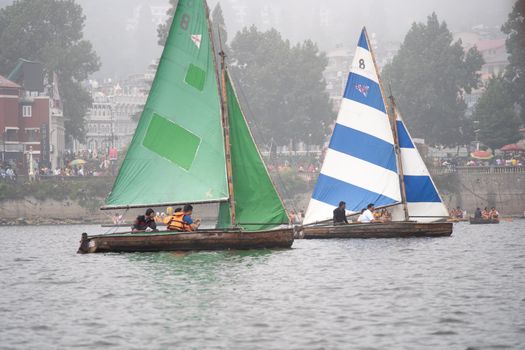 Nanital, Uttarakhand, India - circa 2022 : small colorful sailboat with people sitting on it passing row boats and pedal boats on a foggy day with buildings in the background on the lake of Nainital in uttarakhand