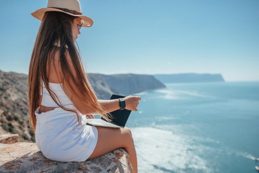 Successful business woman in yellow hat working on laptop by the sea. Pretty lady typing on computer at summer day outdoors. Freelance, travel and holidays concept.