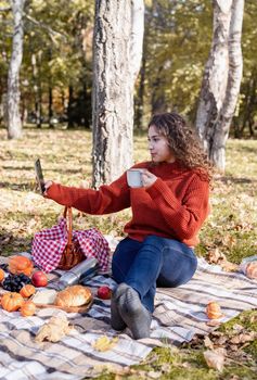 Leisure, free time. Beautiful caucasian woman in red sweater on a picnic outdoors, sitting on a plaid in autumn forest