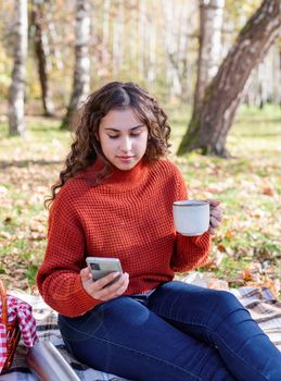 Leisure, free time. Beautiful caucasian woman in red sweater on a picnic outdoors, sitting on a plaid in autumn forest