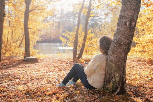 Autumn nature. Young thoughtful woman sitting by the tree in autumn forest