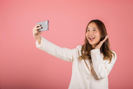 Happy Asian portrait beautiful cute young woman smiling excited  making selfie photo, video call on smartphone studio shot isolated on pink background, female hold mobile phone raise hand say hello