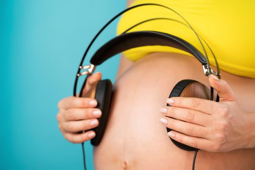 Pregnant woman holding earphones on her belly with baby, enjoying favorite music on blue background. Maternity, motherhood, pregnancy, love concept