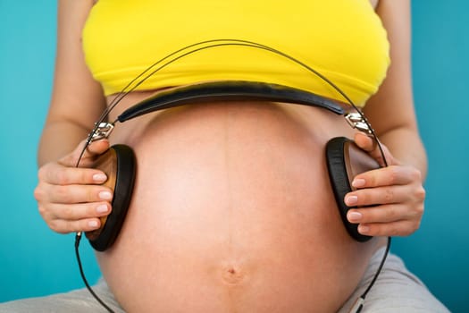Pregnant woman holding earphones on her belly with baby, enjoying favorite music on blue background. Maternity, motherhood, pregnancy, love concept