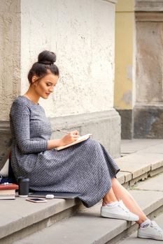 Female student in long dress reading a book on stairs of university building outdoors