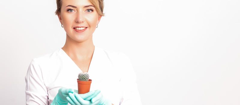 Young beautiful smiling female beautician holding a small cactus in a pot on white background