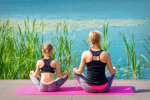 Mother and daughter in sportswear sitting on the mat on the pier near the water outdoor