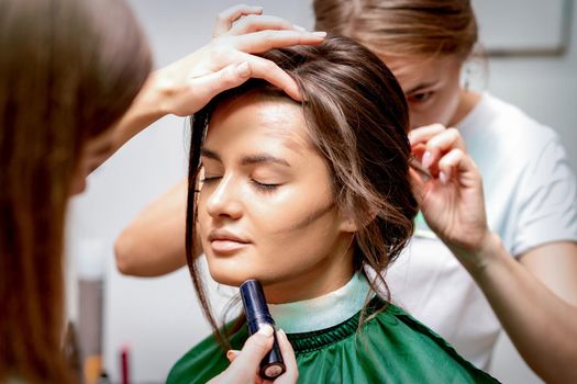 The hairdresser and makeup artist are doing hairstyle and make-up for a beautiful young caucasian woman in a beauty salon