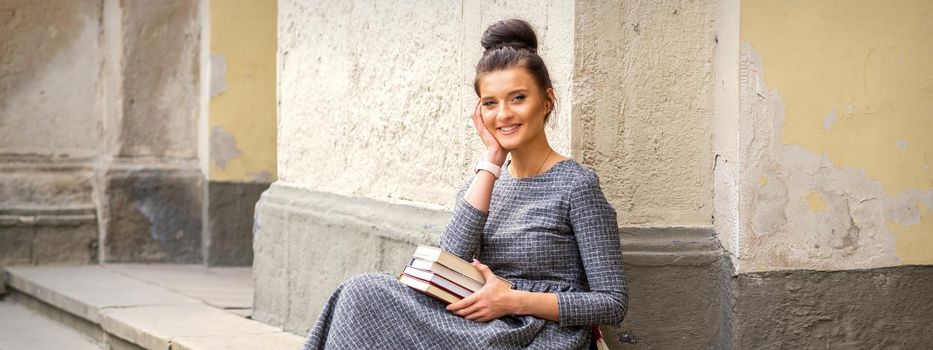 Female student in a long dress holding books looking at camera sitting on stairs of university building outdoors