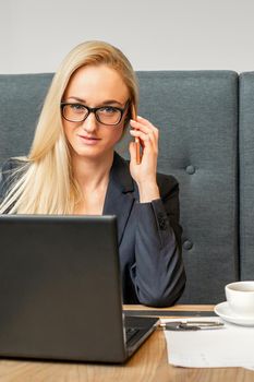 Portrait of a young caucasian businesswoman in glasses calls on a smartphone with a laptop at the table looking at the camera in cafe