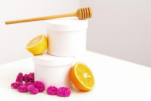 White cosmetic jars with a wooden honey stick, fresh lemon, and chrysanthemums flowers on white background with copy space. Blank space for branding mockup beauty product