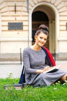Portrait of a beautiful young smiling student studying with a book on the grass near the university building