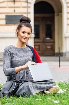 Portrait of a beautiful young smiling student studying with a laptop on the grass near the university building