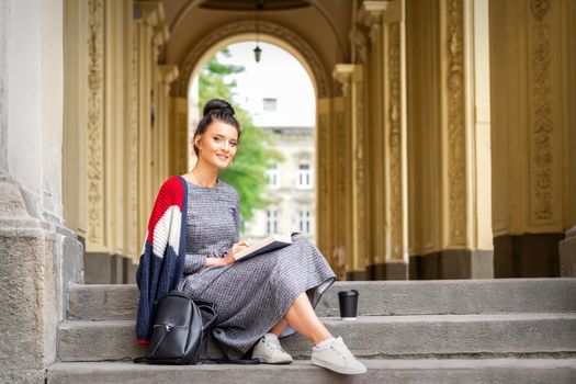 Portrait of a young female student with a book sitting on the stairs of building outdoors