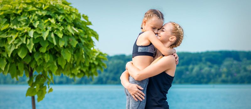 Mother embracing her daughter near the pond wearing sportswear outdoors