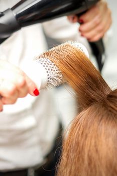 Close up of hairdresser drying long red hair with a hairdryer and round brush in a beauty salon. Selective focus