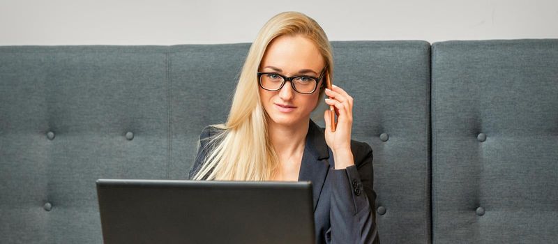 Portrait of a young caucasian businesswoman in glasses calls on a smartphone with a laptop at the table looking at the camera