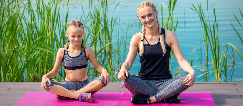Mother and daughter in sportswear sitting on the mat on the pier near the water outdoor