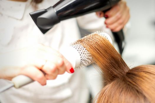 Close up of hairdresser drying long red hair with a hairdryer and round brush in a beauty salon