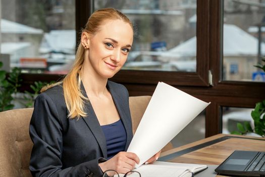 Portrait of a young smiling caucasian businesswoman prepares documents with laptop at the table looking at the camera in cafe