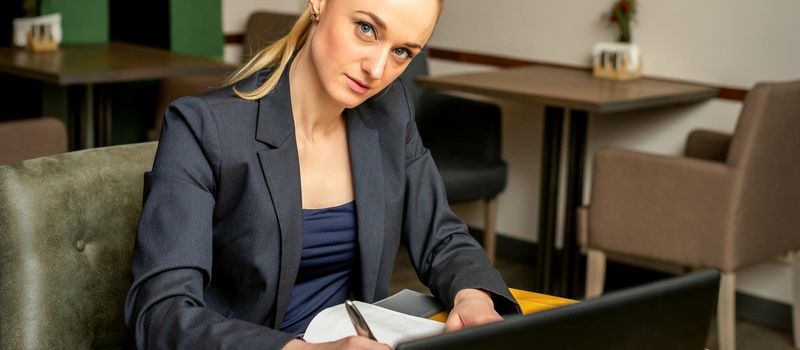 Portrait of a young caucasian businesswoman with pen and laptop at the table looking at the camera in a cafe