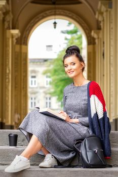 Portrait of a young female student with a book sitting on the stairs of building outdoors
